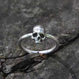 May include: A silver skull ring with a detailed skull design. The ring is on a rough, gray, wooden surface.