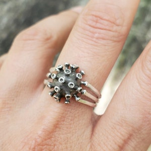 May include: A silver ring featuring a three-dimensional virus design. The ring is worn on a finger.