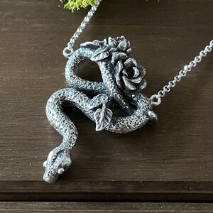 May include: A silver necklace featuring a pendant of a snake entwined around a rose. The snake and rose are intricately detailed and lifelike.
