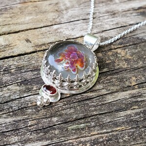 May include: A silver necklace with a round pendant featuring a clear glass dome with a red and purple flower inside. The pendant is surrounded by a silver filigree design and has a small red gemstone hanging from the bottom.