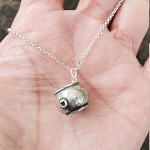 May include: A silver necklace with a small, round, textured pendant shaped like a cauldron. The pendant has a small, round hole in the side.