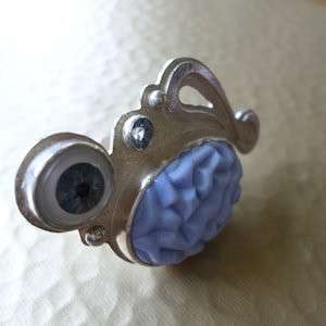 May include: A silver ring with a blue flower design and a glass eye. The ring is made of metal and has a textured surface.