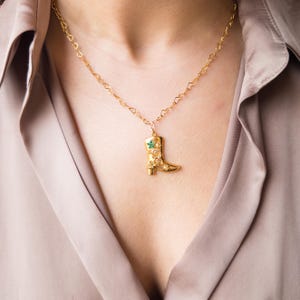Puede incluir: Un collar de oro con un colgante en forma de bota. La bota está decorada con flores verdes y doradas.