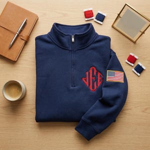 Könnte beinhalten: Marineblaues Sweatshirt mit Viertelreißverschluss, rotem Monogramm und einem amerikanischen Flaggenaufnäher auf dem Ärmel. Das Sweatshirt liegt gefaltet auf einer Holzoberfläche, mit Notizbuch, Stift und Kaffeetasse.