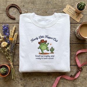 May include: White sweatshirt with the text "Howdy Little Hopper Club" and a cartoon frog wearing a cowboy hat. The frog is next to a cactus. The text "Small but mighty, and ready to boot-scoot!" is below the frog.