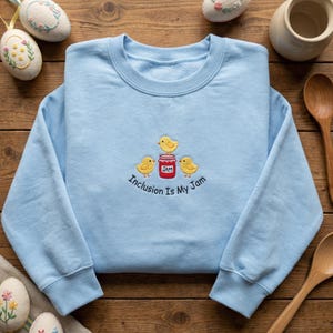 May include: A light blue sweatshirt with an embroidered design featuring three yellow chicks, a red jam jar, and the text "Inclusion Is My Jam". The design is centered on the front of the crew neck sweatshirt.