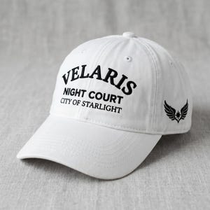 May include: White baseball cap with the text "VELARIS NIGHT COURT CITY OF STARLIGHT" in black. A black wing and star design is on the side. The cap has a curved brim and a button on top.