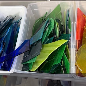 Op de afbeelding: Assortiment van kleurrijke glas-in-lood stukken in doorzichtige plastic bakken. De glasscherven zijn in tinten blauw, groen, geel en rood. Ideaal voor knutsel- of kunstprojecten.