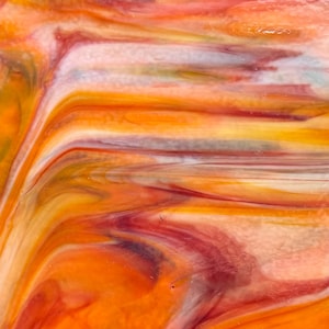Puede incluir: Una lámina de vidrio tintado con un patrón arremolinado y jaspeado de tonos naranja vibrante, rojo intenso y blanco crema. La superficie muestra una textura lisa y brillante con áreas translúcidas y opacas que se mezclan en vetas fluidas.