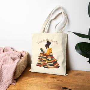 May include: A cream-colored tote bag featuring an illustration of a person reading atop a stack of books. The person is wearing a yellow hoodie. The text "Lost in the Stacks" is arched above the illustration.
