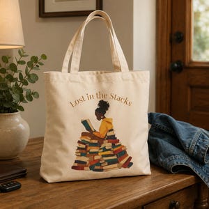 May include: A cream-colored canvas tote bag featuring a graphic illustration of a person with dark skin and a high bun wearing a yellow hoodie, sitting atop a large pile of colorful books. The text "Lost in the Stacks" is printed above the illustration.