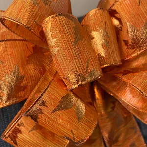 May include: Close-up of a decorative orange bow with gold leaf accents. The bow is made of a textured ribbon with a metallic sheen. The leaves are subtly printed on the ribbon, adding a touch of autumn. Perfect for fall decor.