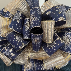 May include: A large decorative bow made of navy blue and gold ribbon. The navy blue ribbon features silver snowflakes, while the gold ribbon has a glittery, striped design. The bow is suitable for holiday or winter decor.