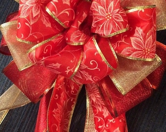 May include: A festive red and gold decorative bow. The bow features layers of red ribbon with a white floral pattern and gold trim. Gold mesh ribbon adds texture. Perfect for holiday decorations or gift wrapping.