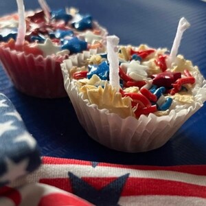 May include: Two red, white, and blue cupcake-shaped candles with white wicks. The candles are decorated with red, white, and blue sprinkles and stars.