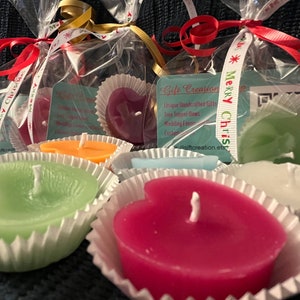May include: Heart-shaped candles in various colors, including pink, green, orange, and white, are displayed in paper cups. Some candles are packaged in clear cellophane bags tied with ribbons that say "Merry Christmas". The image also shows a tag that says "Gift Creation".