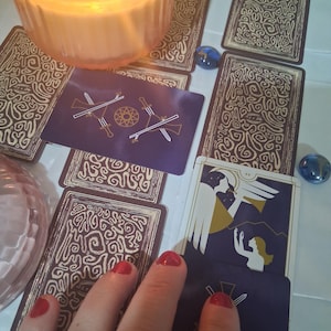 May include: A collection of tarot cards with dark purple backs featuring a gold line-art pattern of two crossed swords and a central star. One card shows an angel blowing a trumpet above a figure. A lit candle and blue glass beads are present.