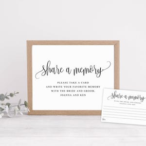 May include: A framed sign and a card with the text "Share a memory" in elegant script. The sign is in a light wood frame, and the card has lines for writing. The text on the sign reads "Please take a card and write your favorite memory with the bride and groom, Joanna and Ken."
