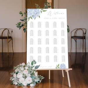 May include: A white wedding seating plan on an easel, decorated with blue hydrangea and greenery. The chart includes the names of the guests and the table assignments. The top of the chart says "Welcome" and the names "Audrey & Justin" and the date.