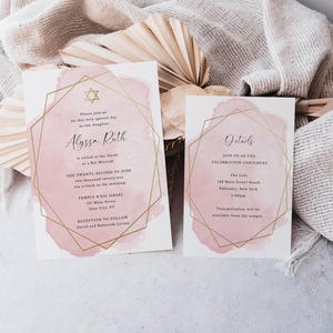 May include: Two white rectangular invitation cards featuring a soft pink watercolour wash background and a thin gold geometric frame. The text includes details for a Bat Mitzvah for Alyssa Ruth on the twenty-second of June at Temple B'nai Israel.