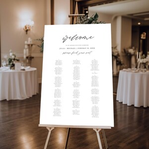 May include: A white wedding reception seating chart on an easel. The sign reads "welcome" in cursive script, followed by the names Julia and Michael, the date February 18, 20XX, and "please find your seat". The names of the guests are listed below.