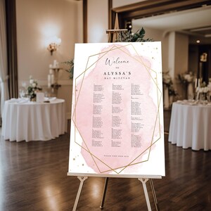 May include: A rectangular seating chart sign on an easel featuring a pale pink watercolor wash background, a gold geometric frame, and black text that reads Welcome to Alyssa's Bat Mitzvah, followed by lists of names and table numbers.