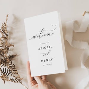 May include: A white wedding welcome card with elegant script lettering. The word "welcome" is at the top, followed by the names "Abigail" and "Henry" and the wedding date. The card is held by a hand.