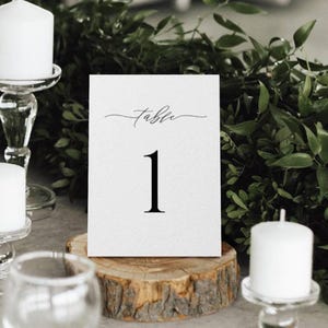 May include: A white table number card with the word "table" in cursive and the number "1" in a bold, black font. The card is displayed on a wooden slice, with candles and greenery in the background. The overall theme is wedding decor.