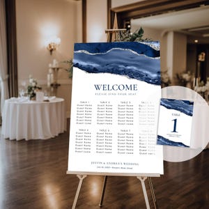 May include: A wedding seating chart on an easel. The sign has a navy blue and silver design at the top with the word "WELCOME" and "PLEASE FIND YOUR SEAT". The names of the guests are listed under the table numbers. The wedding date and names are at the bottom.