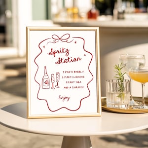 May include: A framed sign with a gold border displays a Spritz Station recipe in red ink. The recipe includes bubbly, liqueur, soda, and a garnish. The sign is on a white table with glasses of drinks in the background.