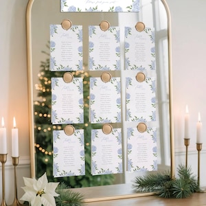 May include: A gold-framed mirror displays nine table assignment cards, each adorned with blue hydrangea floral borders and gold wax seals. The cards are arranged in a grid, with the word "Table" and a number at the top of each.