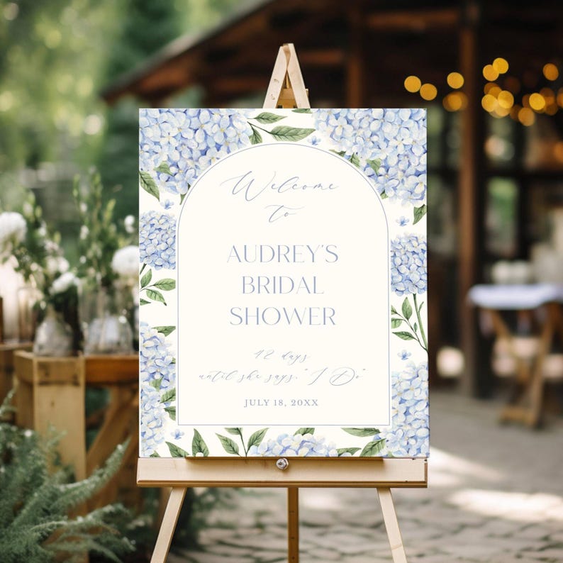 May include: A wooden easel displays a welcome sign for Audrey's bridal shower. The sign features a cream-colored background with a floral border of blue hydrangeas and green leaves. Text includes "Welcome to Audrey's Bridal Shower" and the date.