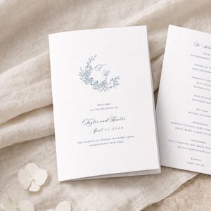 Könnte beinhalten: Weißes Hochzeitsprogramm mit floralem Kranz und den Initialen "T" und "H". Der blaue Text kündigt die Hochzeit von Taylor und Hunter am 11. April 2026 an, an einem Veranstaltungsort in Middletown, New York.