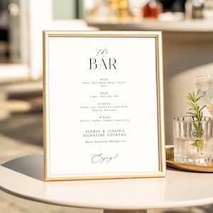 May include: A framed sign with a gold border displays a bar menu. The text reads "The BAR" at the top, followed by lists of wine, beer, spirits, and mixers. The sign also lists a signature cocktail and ends with the word "Enjoy!"
