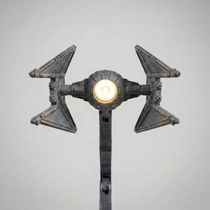 Peut inclure: Lampe grise en forme de chasseur stellaire avec une source de lumière centrale. La lampe présente deux ailes inclinées de chaque côté et une surface texturée détaillée. La lampe est conçue pour ressembler à un chasseur TIE.