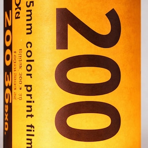 May include: A close-up of a 35mm color print film canister. The canister is black with a yellow label. The label features the number "200" in large brown font, along with the text "35mm color print film".