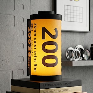 May include: A unique lamp shaped like a 35mm film canister, featuring the number "200" in large, bold text. The lamp has a yellow and black color scheme, with the text "35mm color print film" visible. It sits atop a stack of books.