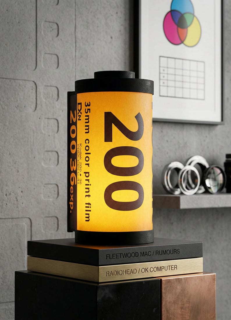 May include: A unique lamp shaped like a 35mm film canister, featuring the number "200" in large, bold text. The lamp has a yellow and black color scheme, with the text "35mm color print film" visible. It sits atop a stack of books.