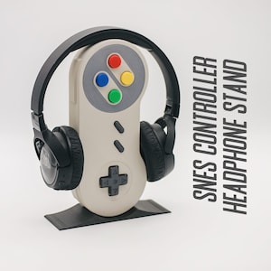 Peut inclure: Un support pour casque noir en forme de manette Super Nintendo Entertainment System (SNES). Le support est en plastique et présente un schéma de couleurs gris et blanc. Le support a une base noire et un support de casque noir de chaque côté.