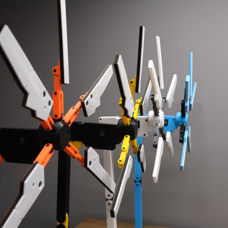 K&ouml;nnte beinhalten: Nahaufnahme von drei bunten, beweglichen, 3D-gedruckten sternf&ouml;rmigen Objekten. Die Objekte bestehen aus schwarzem, wei&szlig;em, orangefarbenem, gelbem und blauem Kunststoff. Die Objekte sind miteinander verbunden und stehen auf einer Holzoberfl&auml;che.