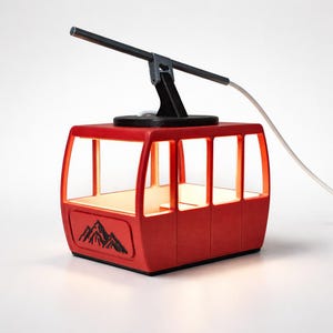 Può includere: Una lampada rossa a forma di cabina di una funivia. La lampada ha una struttura di supporto nera e una barra nera che si estende dalla parte superiore. La cabina presenta una grafica di montagna e un bagliore interno caldo.