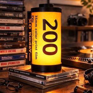 May include: A cylindrical lamp resembling a 35mm color print film canister, with the number 200 prominently displayed. The lamp is yellow and black, sitting atop a stack of CDs and books. A vintage camera and vinyl records are in the background.