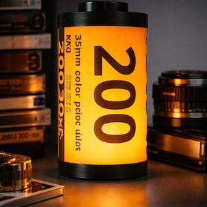 May include: A black and yellow film canister-shaped lamp with the number 200 prominently displayed. The lamp has a warm, inviting glow, and the text "35mm color" is visible. Other photography equipment is in the background.