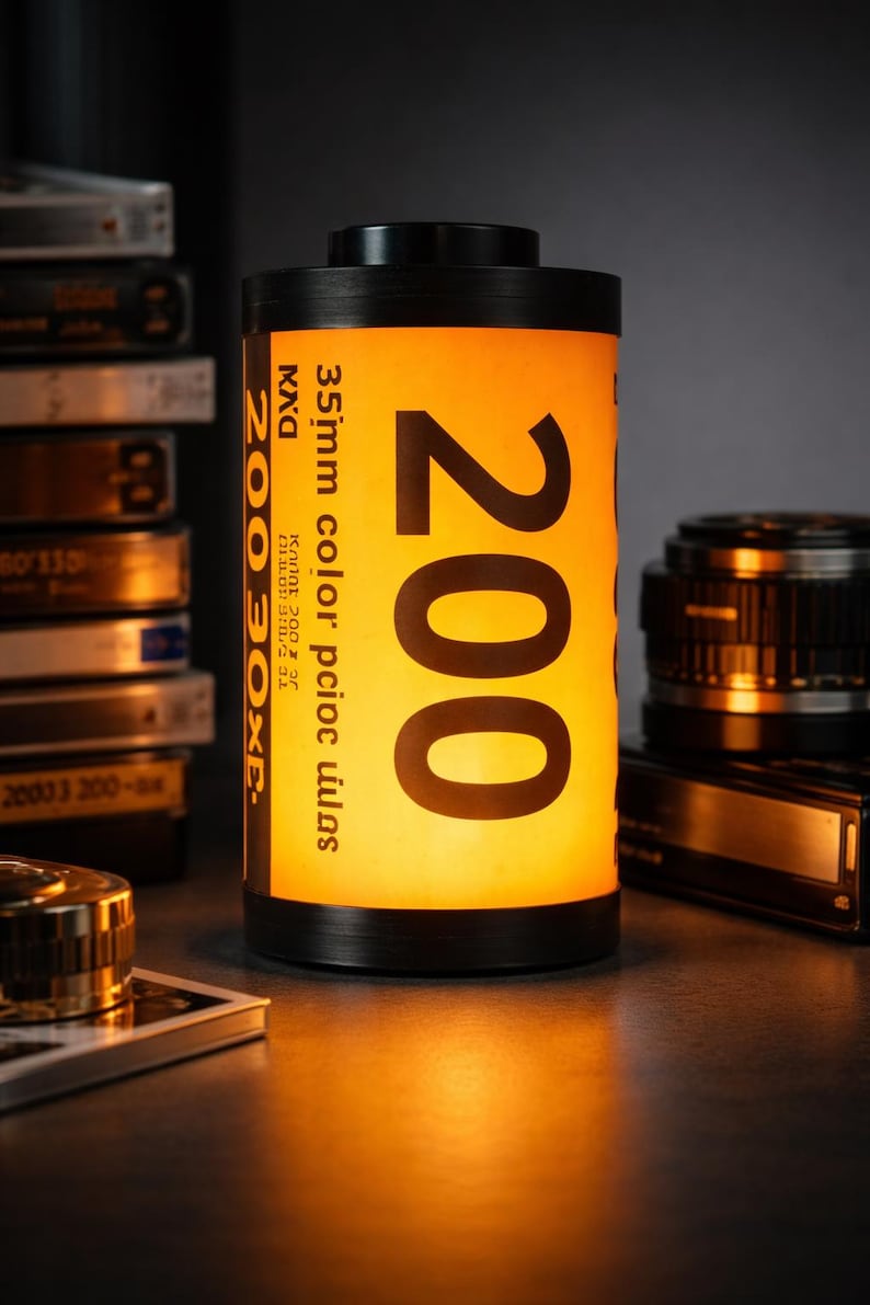 May include: A black and yellow film canister-shaped lamp with the number 200 prominently displayed. The lamp has a warm, inviting glow, and the text "35mm color" is visible. Other photography equipment is in the background.