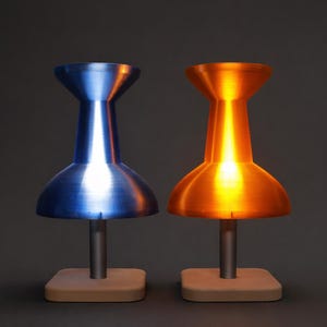 May include: Two pushpin-shaped lamps, one blue and one orange, each with a silver stem and a square, light-coloured base. The lamps are illuminated, casting a warm glow. The background is a dark grey.