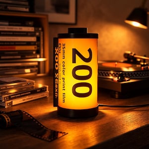 May include: A cylindrical lamp designed to resemble a 35mm film canister. The lamp has a black base and top, with a bright yellow center displaying the number "200" and text "35mm color print film". The lamp is on a wooden surface.