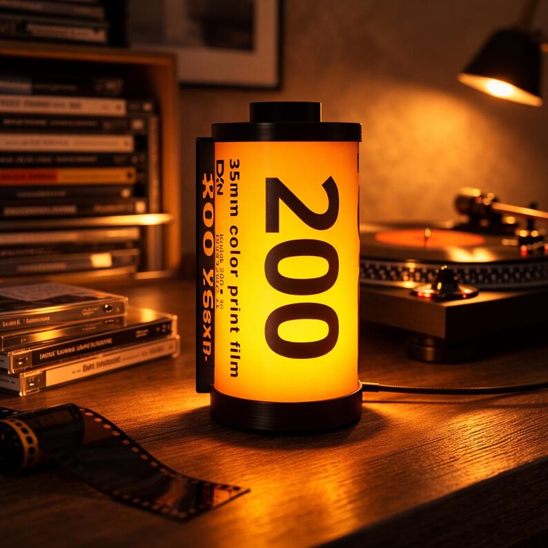 May include: A cylindrical lamp designed to resemble a 35mm film canister. The lamp has a black base and top, with a bright yellow center displaying the number "200" and text "35mm color print film". The lamp is on a wooden surface.
