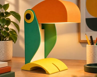 Op de afbeelding: Een toekanvormige tafellamp met een grote oranje snavel, een groen lichaam en gele voeten. De lamp geeft warm licht onder de snavel. Het ontwerp bestaat uit platte, effen gekleurde panelen met een zwart cirkelvormig accent aan de zijkant.