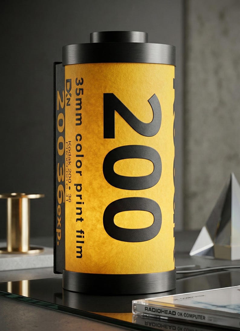 May include: A cylindrical lamp designed to resemble a 35mm color print film canister. The lamp features a yellow and black design with the number "200" prominently displayed. The text "35mm color print film" is also visible. The lamp is a unique decorative item.