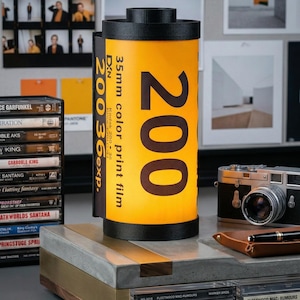 May include: A decorative lamp shaped like a 35mm Kodak film canister, with the number "200" prominently displayed. The lamp has a black top and base, and the film canister is yellow with black text. A vintage camera and cassette tapes are also visible.